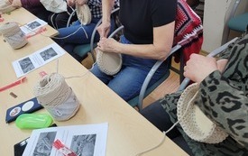 Przy stoliku osoby wykonujące koszyki na szydełku. Na stole wydruki, sznurki bawełniane.