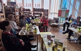 Kobiety zebrane na spotkaniu. Na stolikach wiosenne kwiaty w doniczkach, filiżanki, talerzyki ze słodkościami.