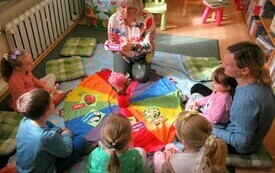Na dywanie przy kolorowej chuście siedzi grupa dzieci. Na środku hiacynt. 