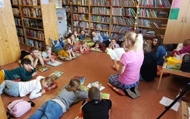 Grupa dzieci siedzi na podłodze i słucha kobiety czytającej książkę.