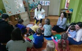 Bibliotekarka pokazuje książkę dzieciom i dorosłym, którzy siedzą na dywanie.