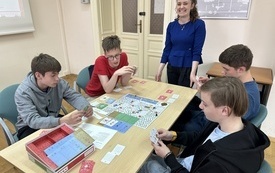 Bibliotekarka stoi obok stołu, przy którym gra 4 chłopców.