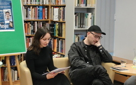 Młody mężczyzna i kobieta siedzą na sofie. Za nimi regały z książkami. 