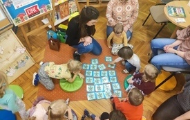 Na dywanie rozłożone karty, dzieci siedzą dookoła wraz z dorosłymi i odsłaniają karty. 