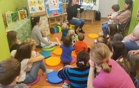 Bibliotekarka czyta książkę i pokazuje ilustracje, wok&oacute;ł dorośli i dzieci. 