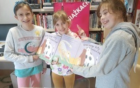 Trzy dziewczynki trzymają książkę. W tle plakat Dyskusyjnych Klub&oacute;w Książki. 