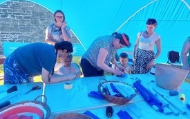 Niebieski namiot a w nim kobiety i dzieci tworzące prace plastyczne. 