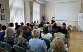 Grupa osób w różnym wieku siedzi i słuicha wystapienia prelegentki