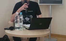 Mężczyzna przy stoliku z laptopem, mówi do mikrofonu.