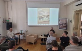 Prelegent i prowadzący spotkanie siedzą na fotelach zwróceni do widowni.