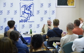 Gość spotkania przemawia do mikrofonu, na pierwszym planie publiczność.