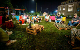 Wieczór, grupa osób siedzi na leżakach, jedna z osób czyta. Na środku stolik z palet