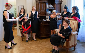 Grupa kobiet w sukienakch stoi, dwie siedzą w fotelach, jedna trzyma kartki papieru.