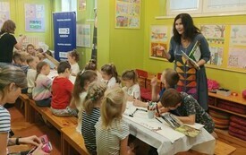 Kobieta z książką stoi przy stole, przy kt&oacute;rym siedzą rysujące dzieci