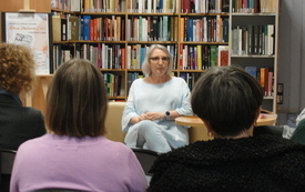 Starsza kobieta siedzi na kanapie, w tle za nią półka z książkami