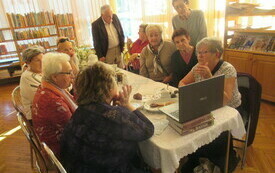 Przy stole kilkoro seniorów, jedna z kobiet pokazuje zdjęcia na laptopie i opowiada.