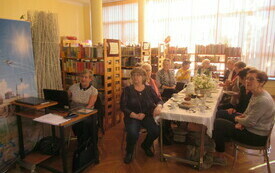 Grupa seniorów przy stole nakrytym obrusem, oraz zastawą i słodkościami. Jedna kobieta siedzi przy biurku przy komputerze.