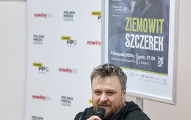 Pisarz w bluzie z kapturem m&oacute;wi do mikrofonu, za nim materiały reklamowe.