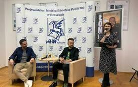 Łona i Adam Bienias siedzą przy stole, przy nich stoi kobieta i m&oacute;wi do mikrofonu. 