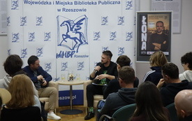 Adam Bienias m&oacute;wi do mikrofonu, obok siedzi łona, w tle plakat z Łoną i logotypami DKK i Podkarpackie. 