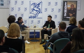 W centrum siedzą Łona, raper i Adam Bienias, dziennikarz, przed nimi grupa ludzi siedzących na krzesłach. W tle logo WiMBP - pegaz. 