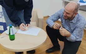 Przy ogkrągłym stoliku mężczyzna i młoda dziewczyna, dziewczyna trzyma w ręku jajko. 