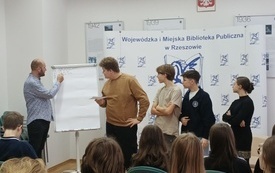 Mężczyzna pisze na tablicy obok stoją młodzi ludzie, chłopcy i dziewczyna.