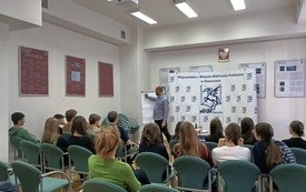 Mężczyzna pisz na tablicy, przed nim zgromadzona publiczność.
