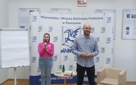 Kobieta w r&oacute;żowym sweterku m&oacute;wi przez mikrofon, obok stoi mężczyzna z zarostem w niebieskiej koszuli.