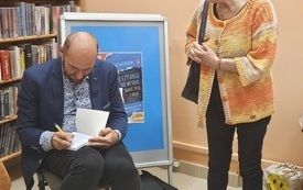 Mężczyzna wpisuje dedykację do książki, obok kobieta w pomarańczowym sweterku.