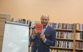 Mężczyzna trzyma w ręku książkę &quot;Dlaczego warto czytać&quot;. W tle regały z książkami.