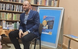 Mężczyzna siedzi na krześle, za nim regał z książkami i potykacz z plakatem zapowiadającym spotkanie. 