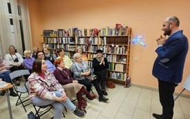 Mężczyzna w marynarce stoi przed grupą siedzących kobiet. Obok kobiet regał z książkami.