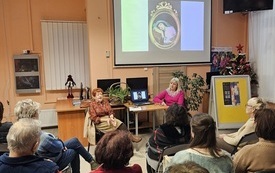 Na ścianie rzutnik z prezentacją, obrazu. Dwie kobiety pośrodku sali zwr&oacute;cone do widowni. 