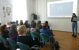 Młoda kobieta z książką w rku stoi pośrodku sali i przemawia. Widownia się przysłuchuje.