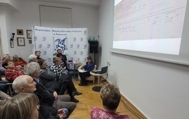 Widownia zebrana na spotkaniu. Prowadząca siedzi i czyta.