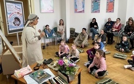 Grupa dzieci siedzi na podłodze w bibliotece słuchając opowieści kobiety w słowiańskim stroju