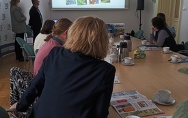 Widok na salę i bibliotekarki siedzące przy stole. Na ścianie prezentacja.