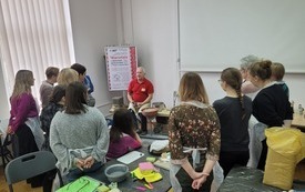 Starszy mężczyzna siedzi przy kole garncarskim, obok grupa kobiet.