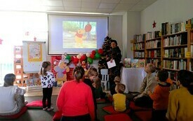 Na dywanie w bibliotece dzieci wraz z opiekunami, w tle prezentacja multimedialna.