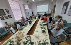 Duży st&oacute;ł z materiałami florystycznymi, przy kt&oacute;rym siedzą kobiety w r&oacute;żnym wieku.