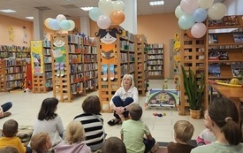 Bibliotekarka siedząca na podłodze przed regałami, zwrócona do dzieci.