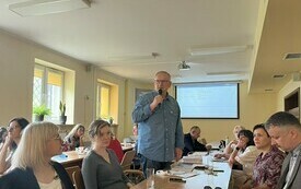 Mężczyzna w niebieskiej koszuli przemawia do współuczestników spotkania.