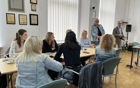 Fotograf prowadzący szkolenie stoi przy stole, przy kt&oacute;rym siedzą uczestniczki. 