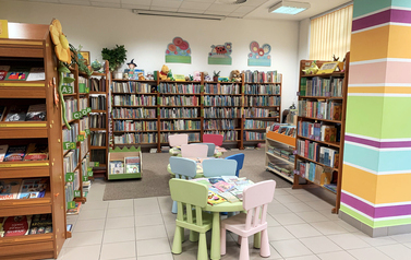 Sala z regałami z książkami, na środku kolorowe stoliki i krzesełka dla dzieci