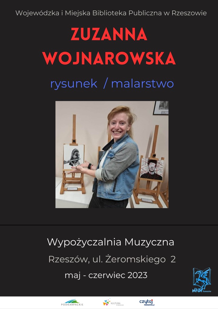 Uśmiechnięta dziewczyna w blond włosach stoi obok sztalug z pracami graficznymi.